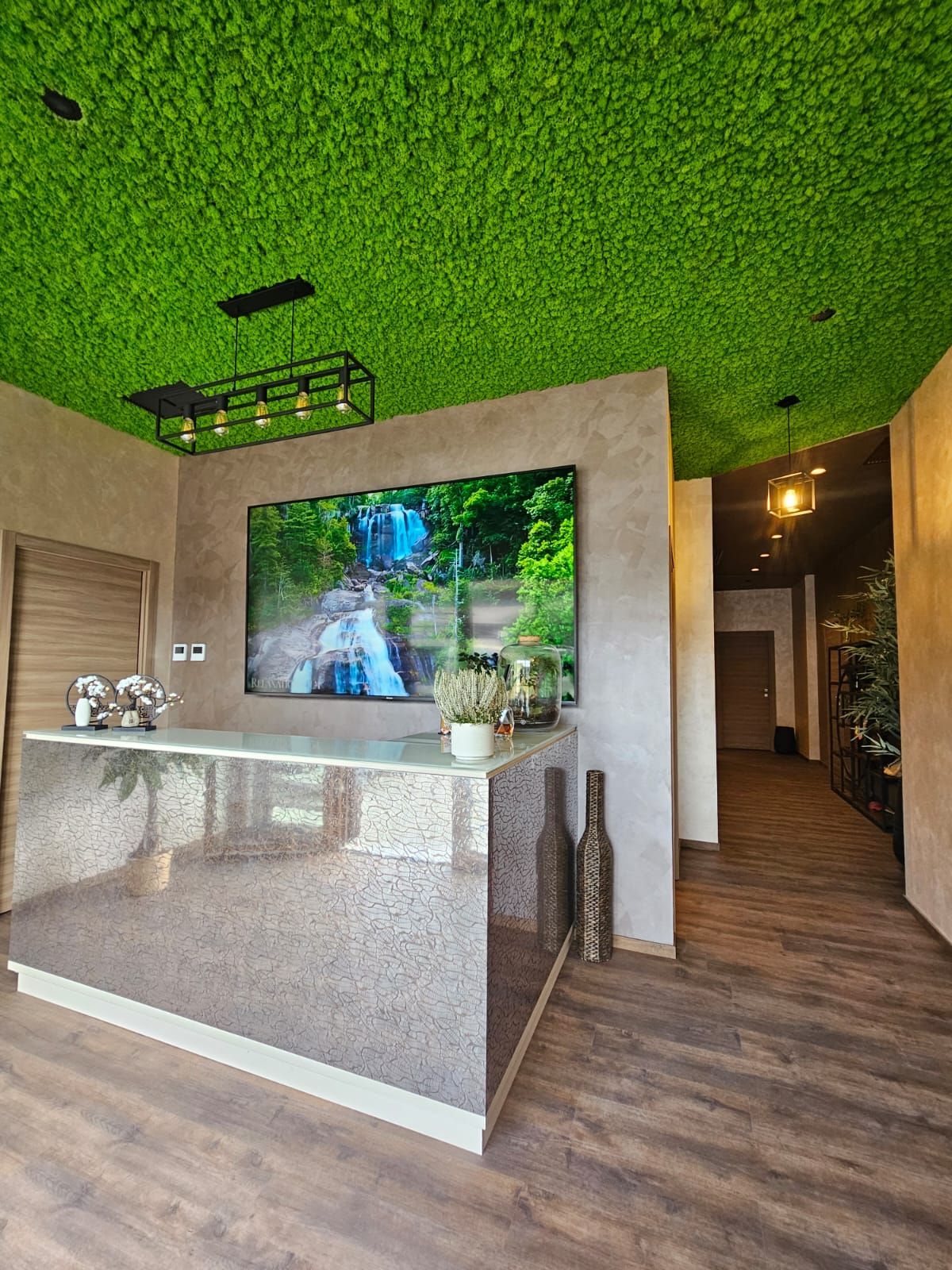 Banco della reception con immagine di cascata, soffitto in muschio e pavimento in legno dai toni caldi.