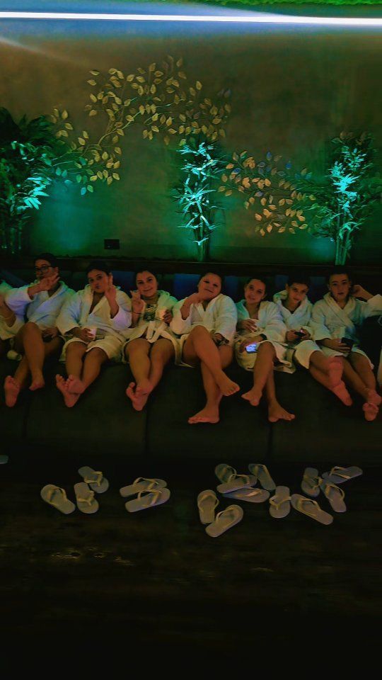 Persone in accappatoio sedute su un divano scuro, con infradito davanti. Luci verdi e fogliame sullo sfondo.