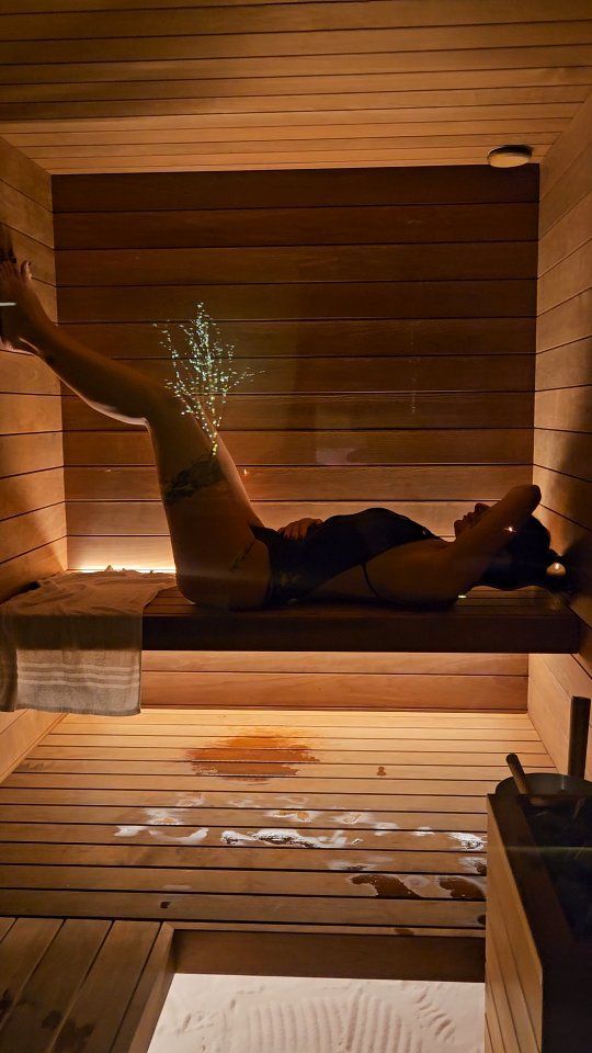 Una donna è sdraiata in una sauna di legno, con le gambe sollevate. La luce illumina l'interno.