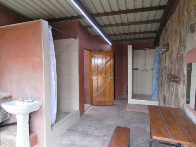 Outdoor shower room with showers, sinks, wooden door, and table; earth tone colors.