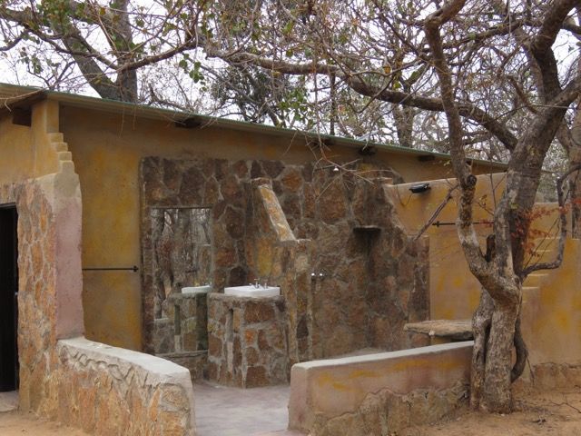 Outdoor bathroom with stone walls, tan plaster, and a large mirror. A tree is nearby.