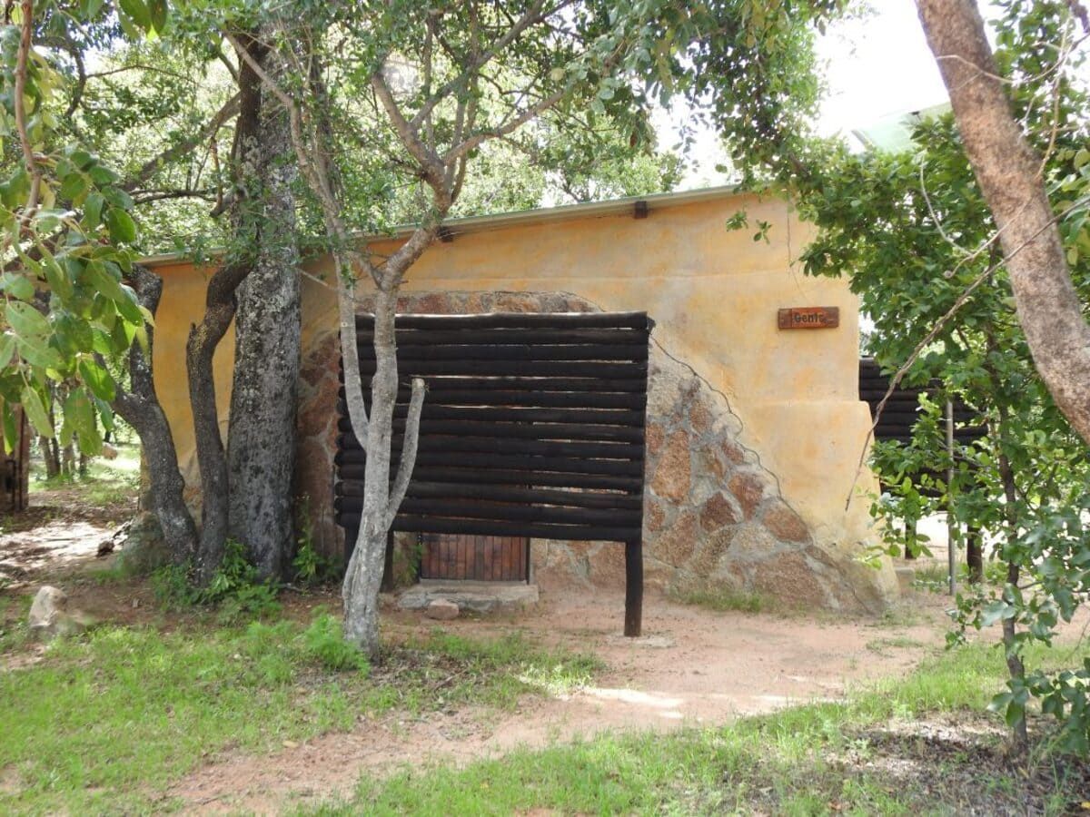 Small, yellow-walled building with a dark wooden sign, partially obscured by trees, in a grassy outdoor area.