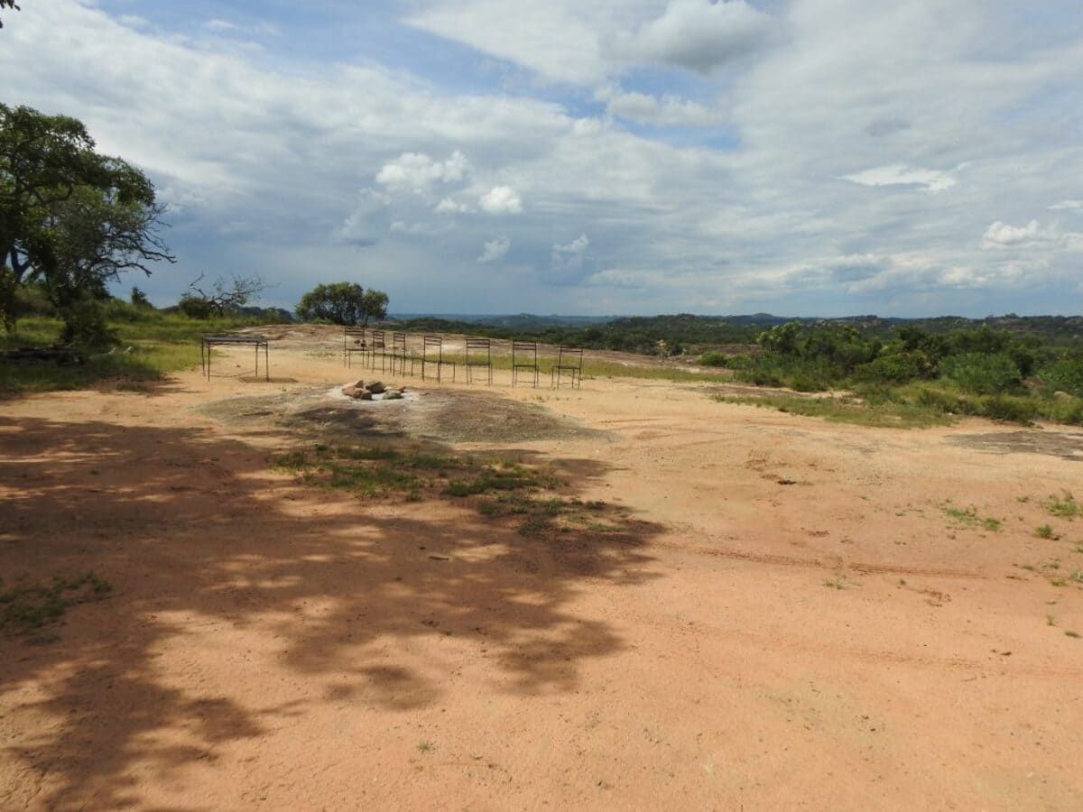 Dirt field under cloudy sky with sparse vegetation and wooden structures.