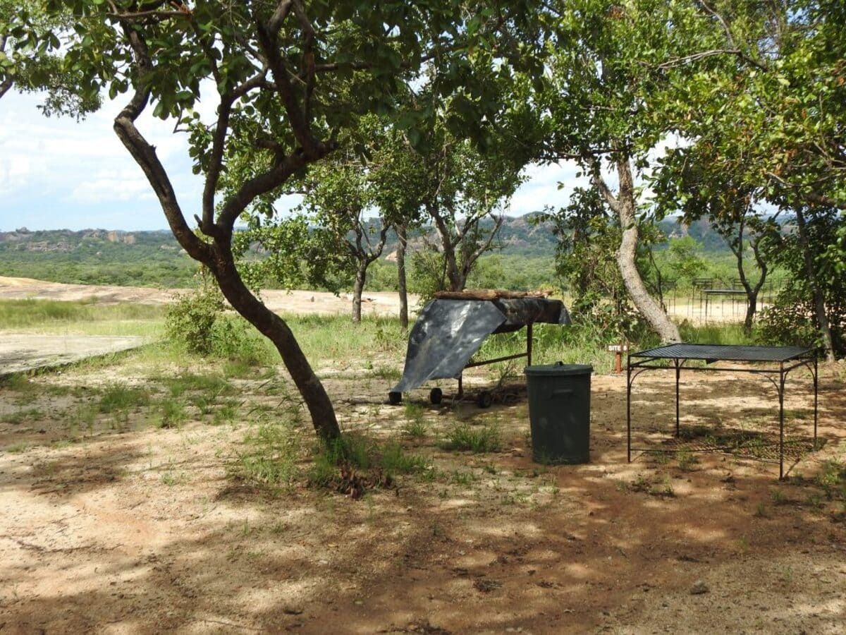 A shaded campsite in a dry landscape, with a grill, table, and trash can near a sparse tree.