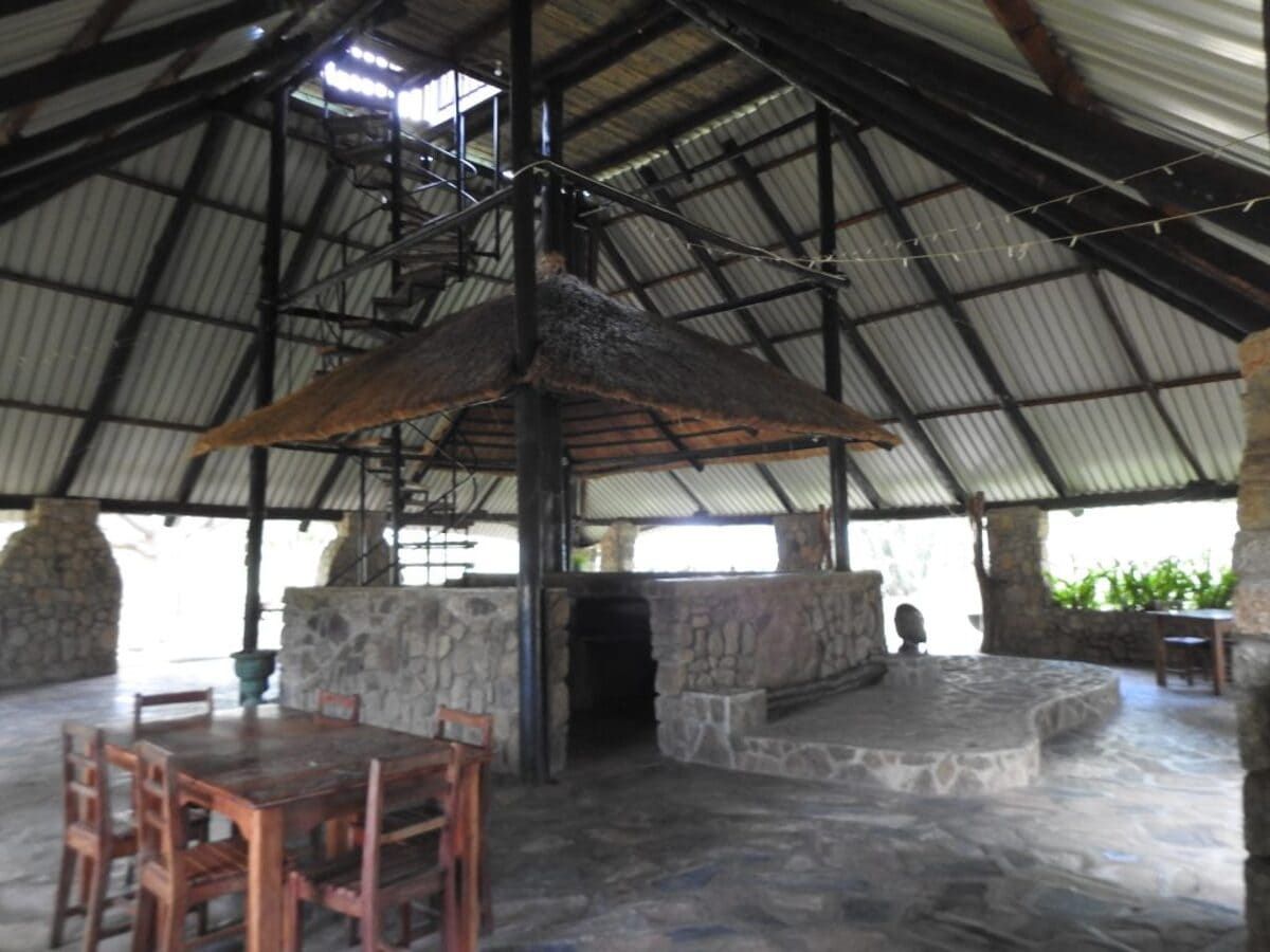 Open-air dining area with a central fireplace, stone pillars, thatched roof, spiral staircase, and wooden furniture.