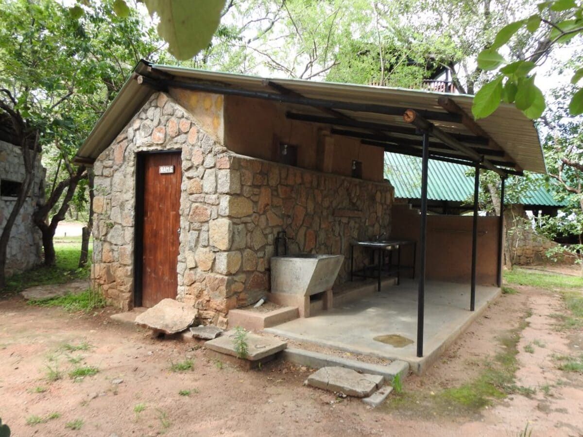 Stone building with brown door, open-air counter, and metal roof in a shaded outdoor area.