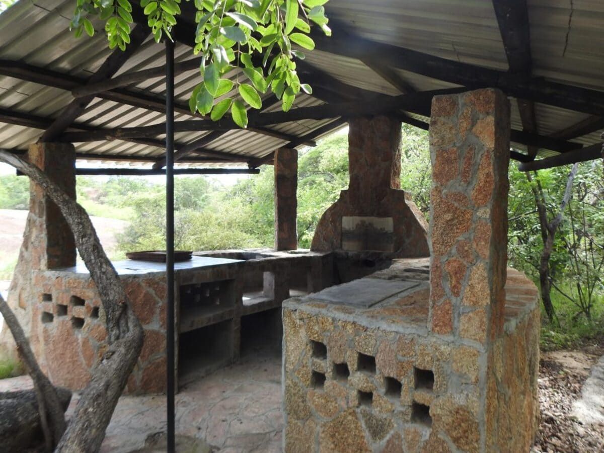 Stone and wood picnic shelter with a grill and counter, set in nature.