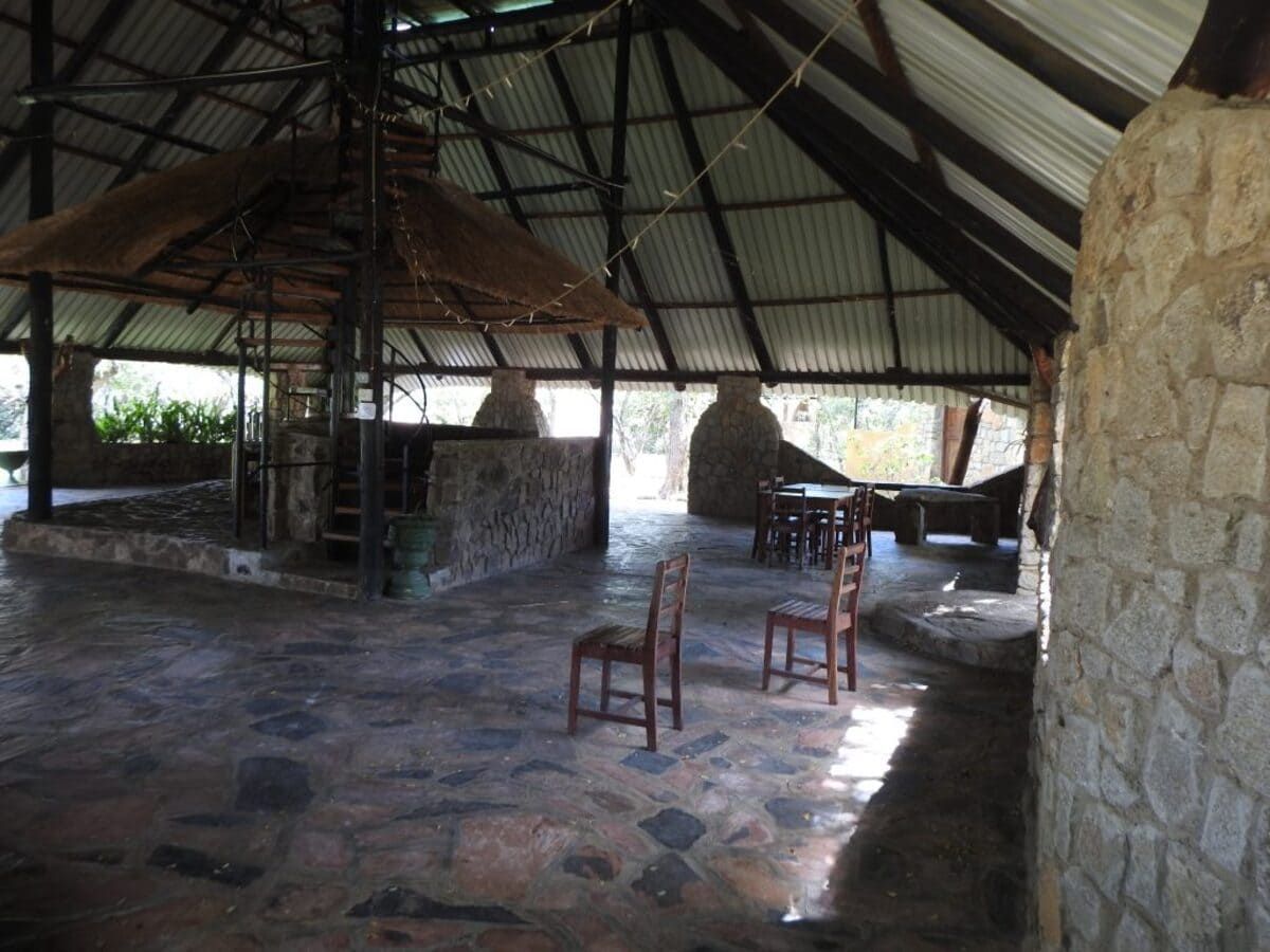 Open-air structure with stone walls and tiled floor. Wooden chairs sit on the floor.