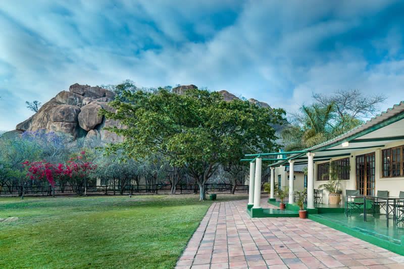 Lush green yard with a brick path leading to a white building with a green trim, against a backdrop of a rocky mountain.