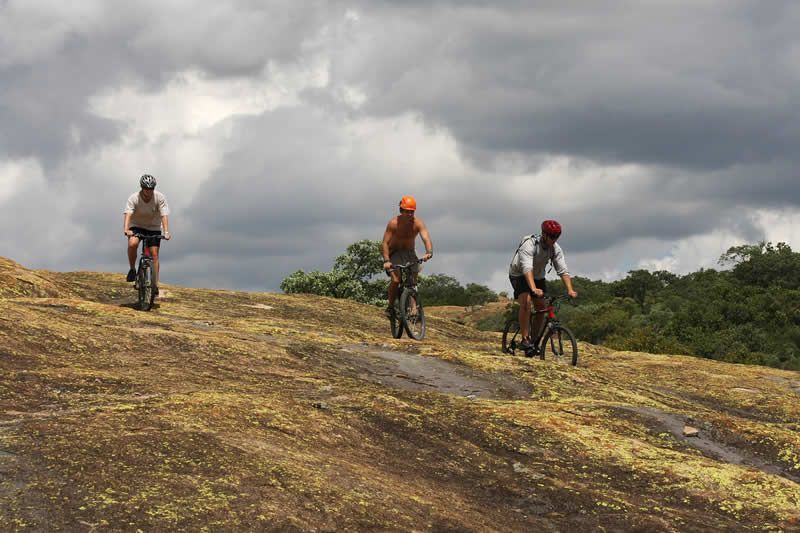Three cyclists riding bikes on a rocky, sloped terrain under a cloudy sky.
