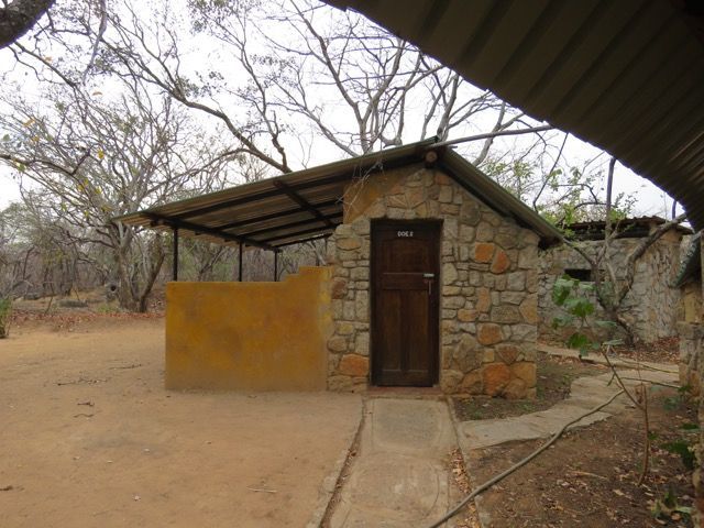 Stone building with a corrugated roof, brown door, and yellow wall in a wooded area.
