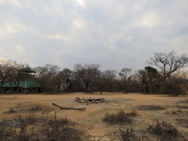 A dry, open landscape with trees, a building with a green roof, and a cloudy sky.