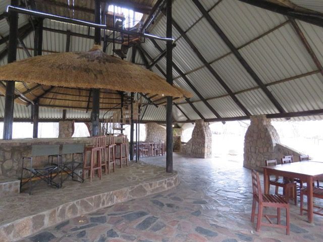 Open-air bar and dining area with thatched roof, stone walls, wooden furniture, and bar stools.