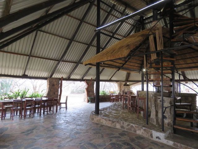 Open-air dining area with a metal roof, stone floor, and wooden tables/chairs. A spiral staircase leads up to a thatch roof.