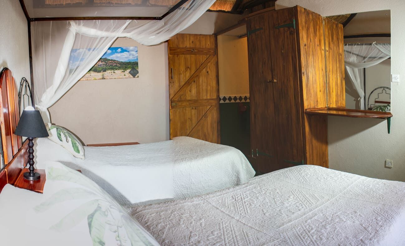 Two beds in a rustic room with wooden features, mosquito net, lamp, and a mirror.