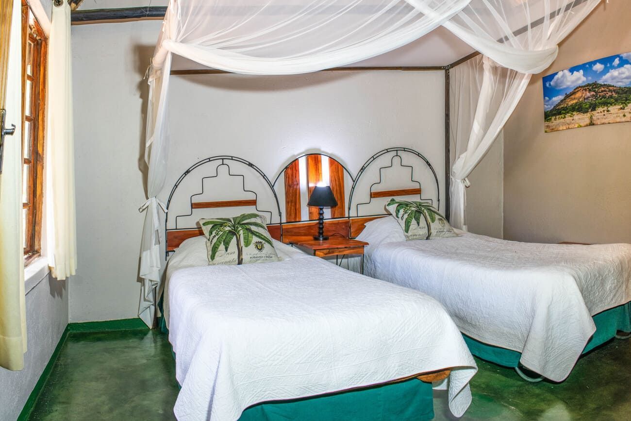 Twin beds in a bedroom with mosquito netting and decorative headboards, a window, and a landscape photo.