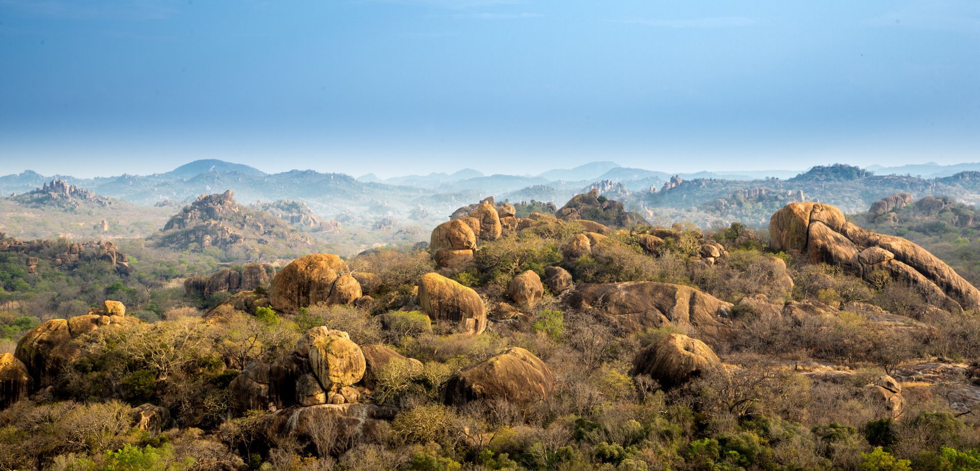Hilly landscape under a blue sky, with trees and rock formations in the foreground.