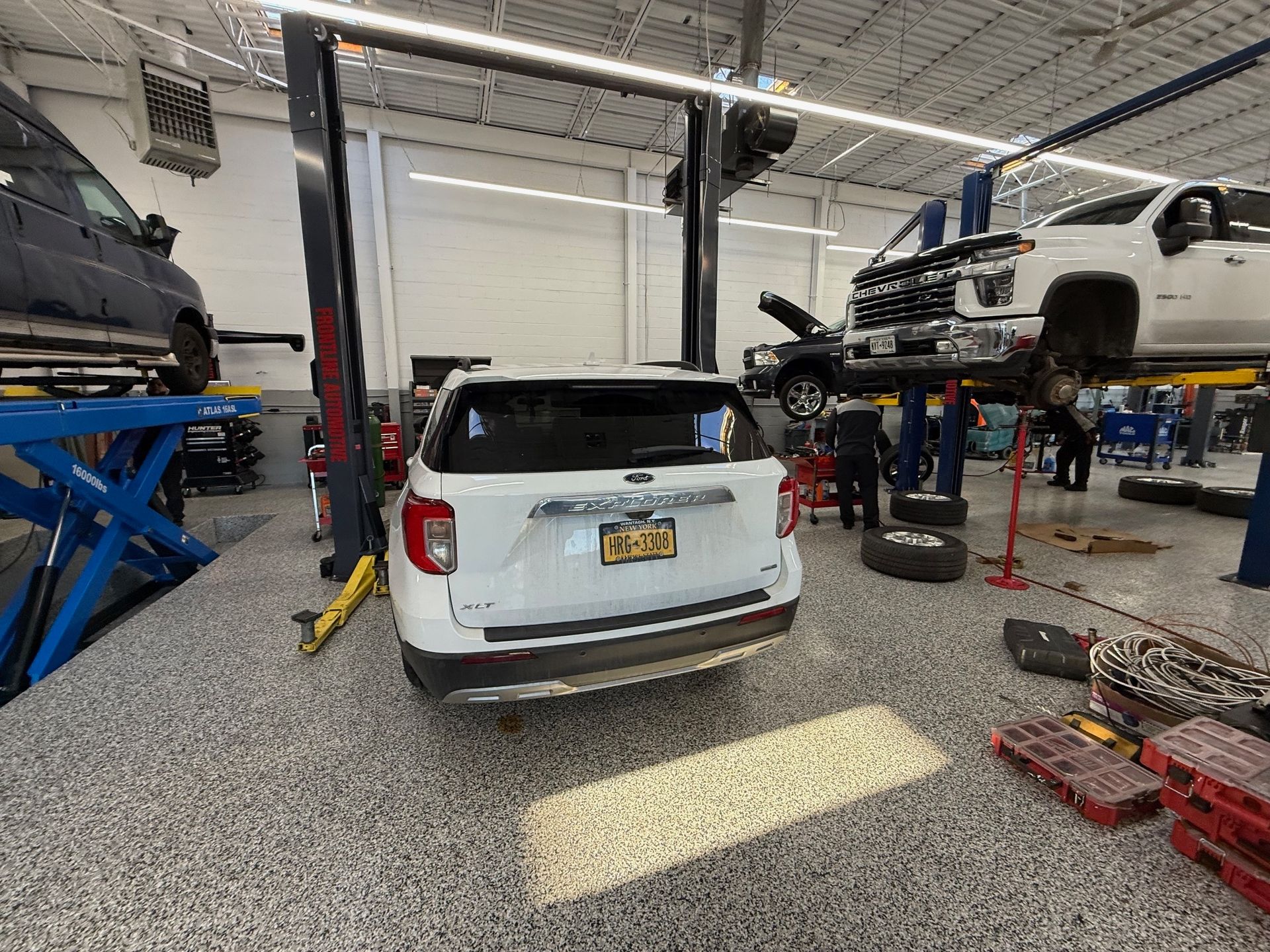 A white SUV parked in a brightly lit automotive repair shop with other vehicles being serviced on lifts.