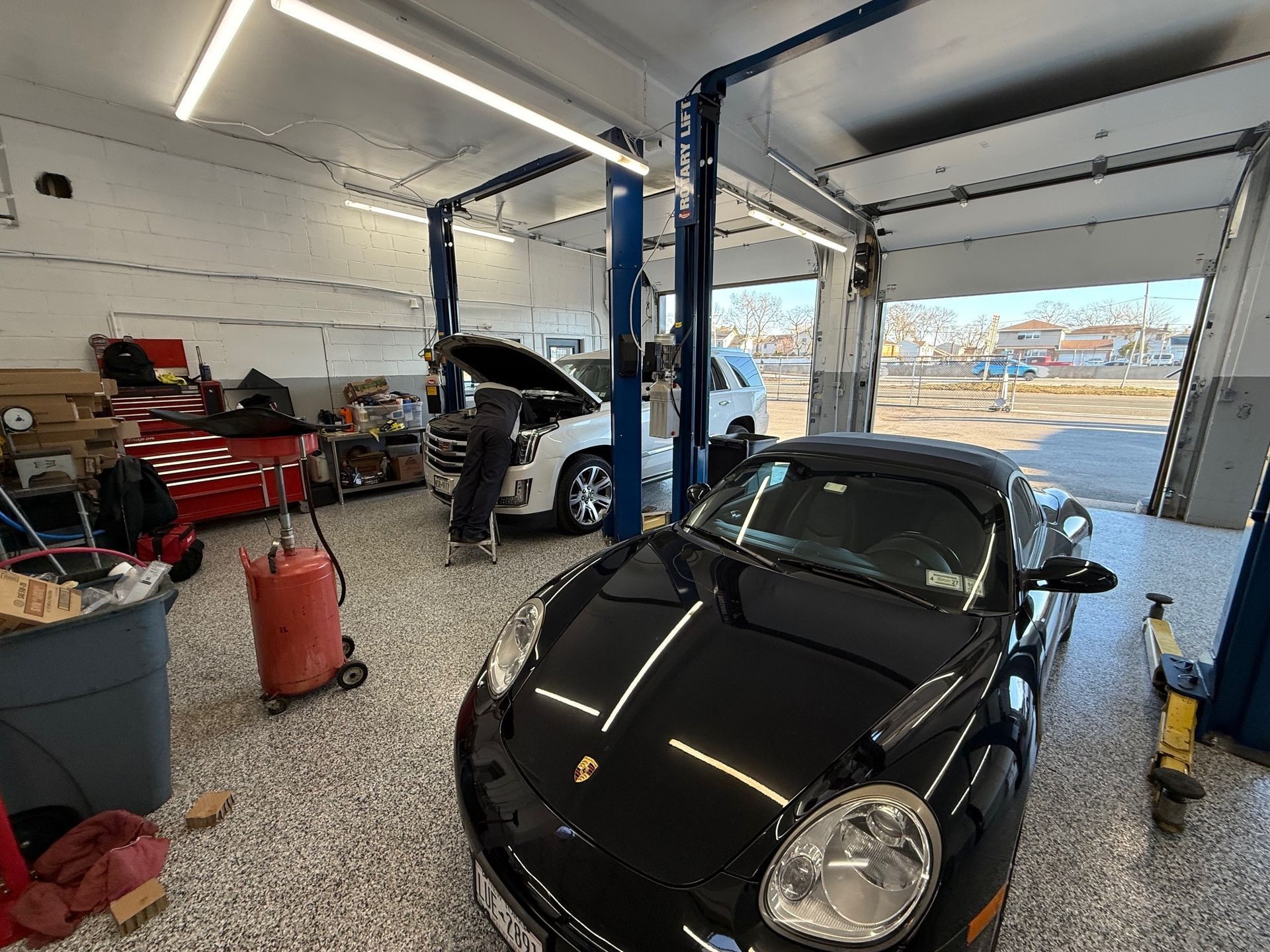An auto repair shop featuring a black sports car in the foreground and a technician working on a white SUV in the back.