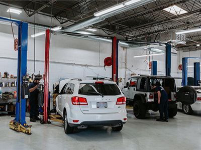 Mechanics working on vehicles inside a well-lit auto repair shop with lifts; white SUV and jeep in focus. | Frontline Auto Repair