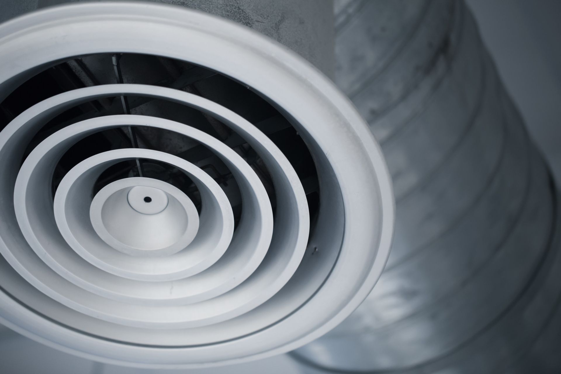 A white circular ceiling air diffuser vent connected to a silver industrial duct.