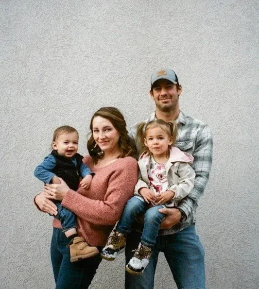 A family of four stands against a textured gray wall, smiling at the camera as the parents hold their two children.