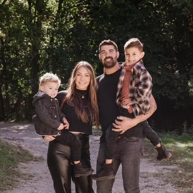A smiling family of four standing together on a sunlit path, surrounded by trees.