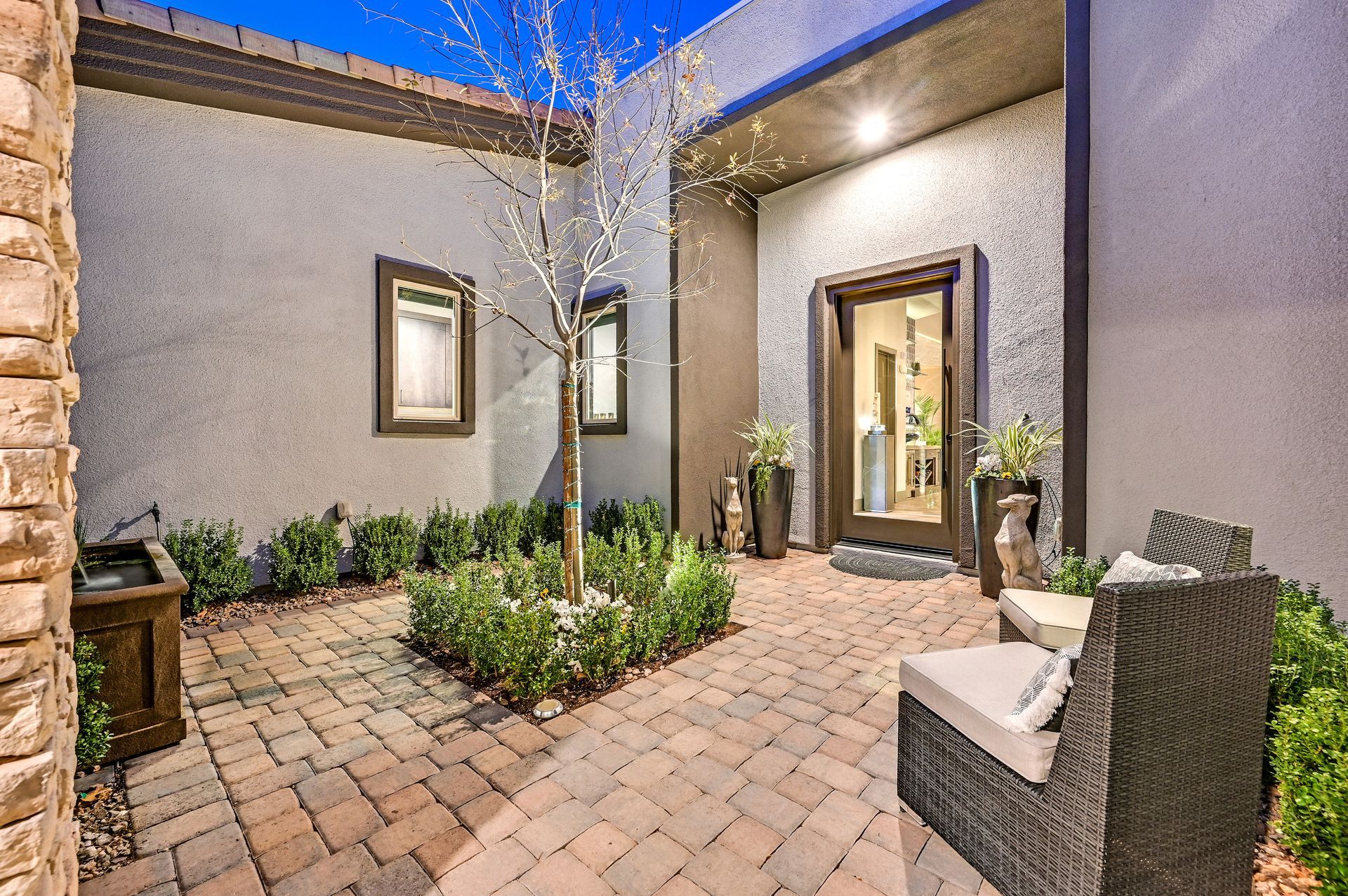 Courtyard with brick pavers, small tree, hedges, and two chairs near front door.
