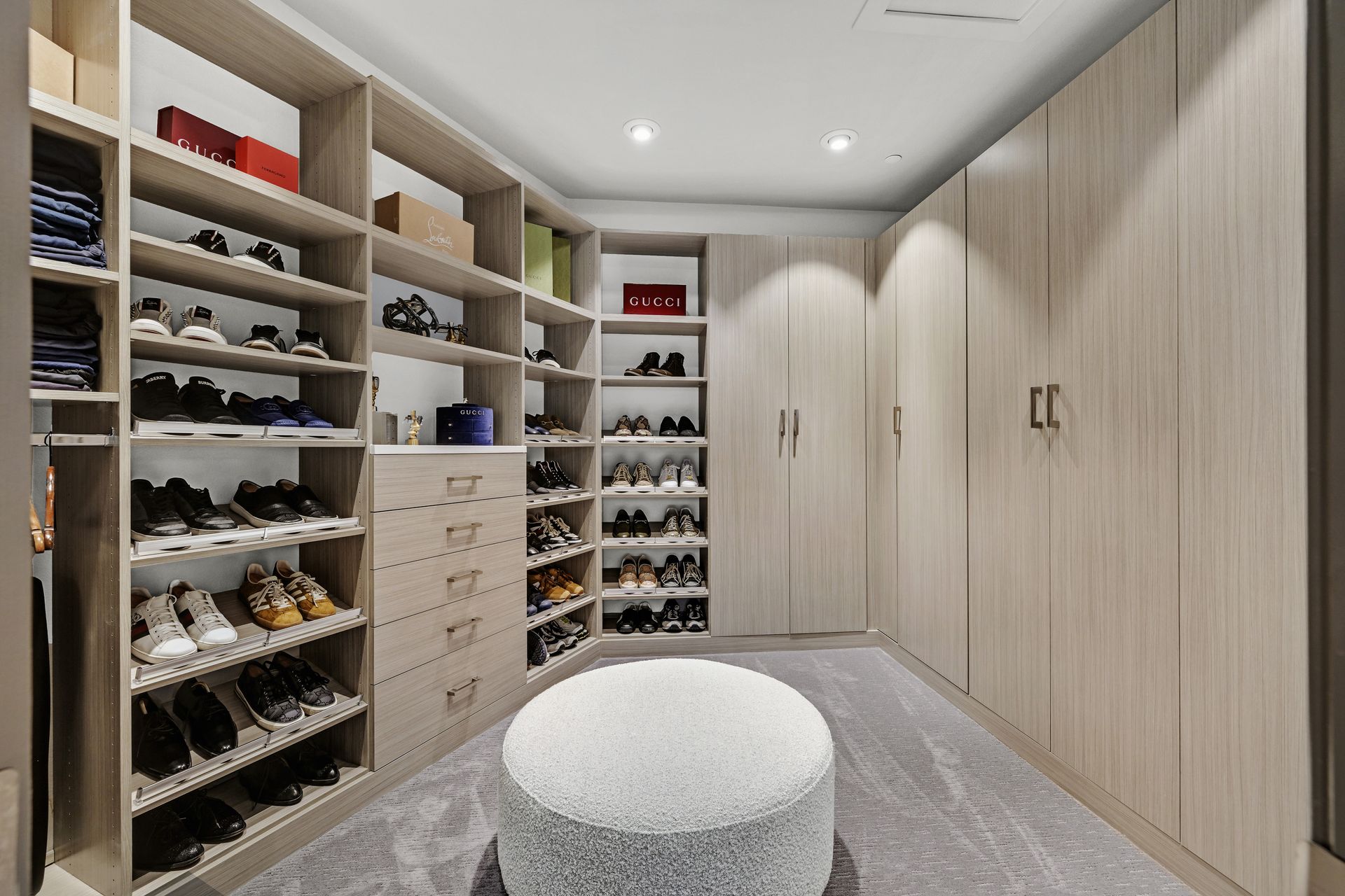 Walk-in closet with shelves and drawers filled with shoes and storage boxes; a cushioned ottoman is in the center.