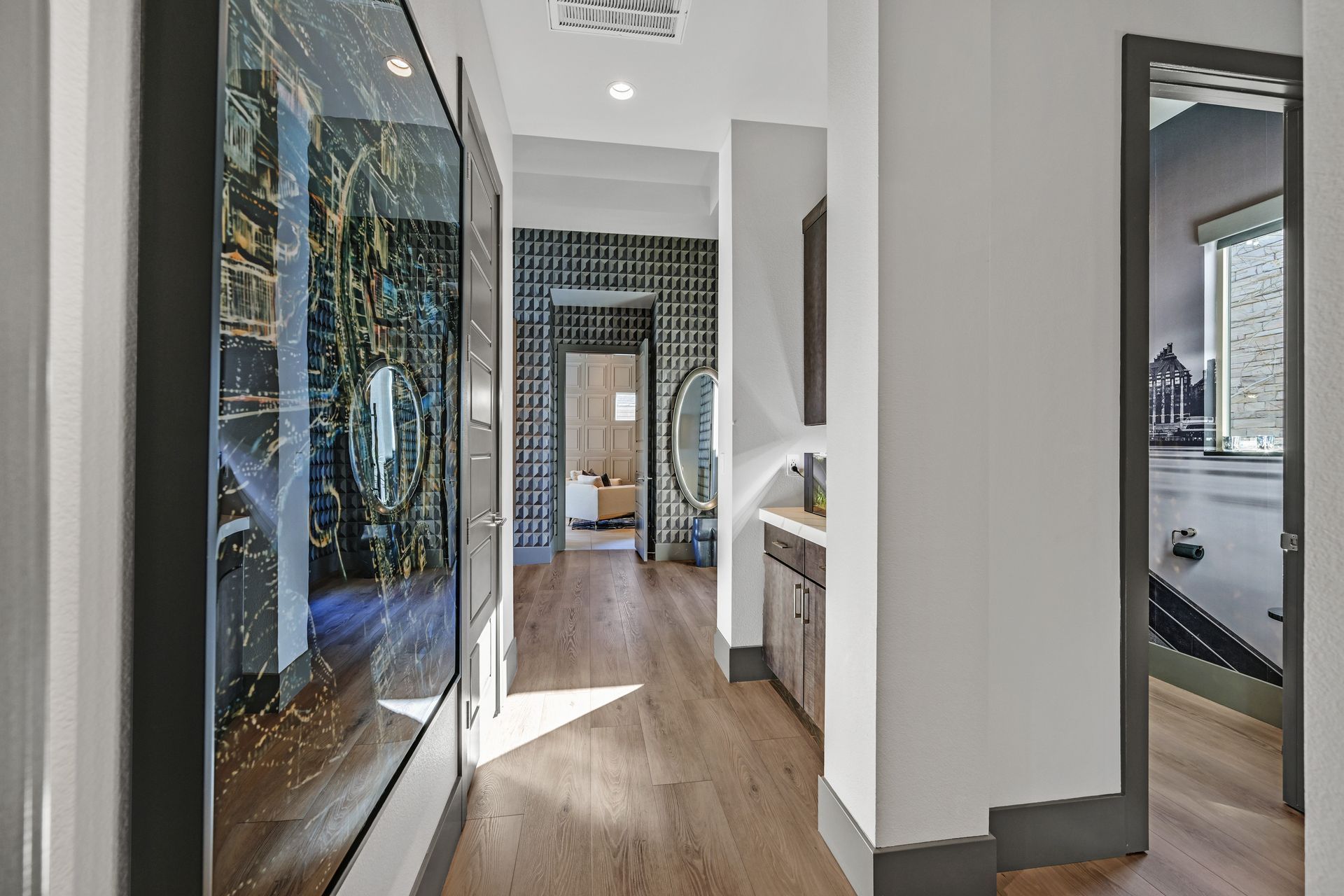 Hallway with light wood floors, gray walls, and artwork. A room is visible at the end of the hall.