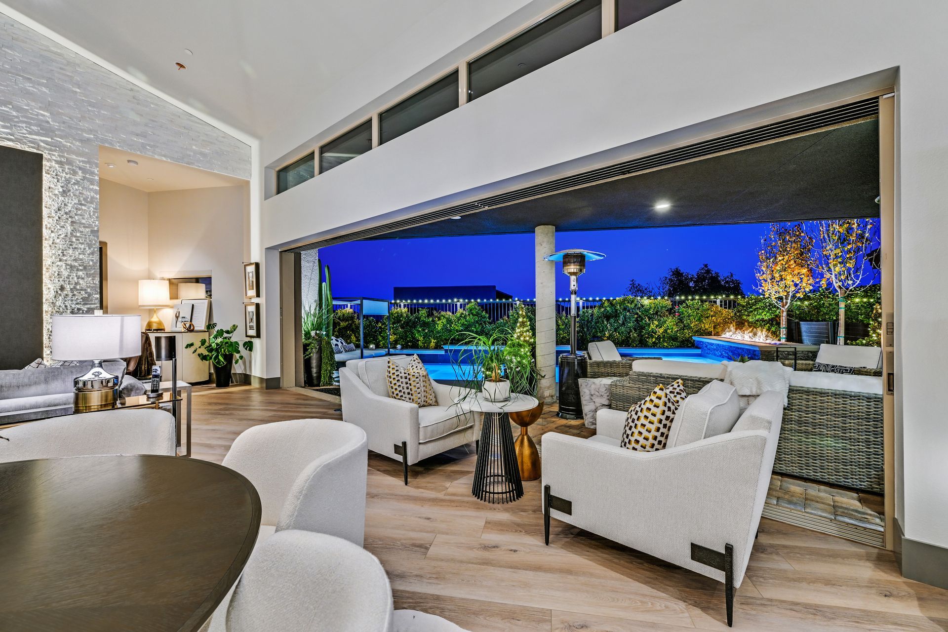Open-air luxury living room with a nighttime view of a pool. White chairs and couches, wooden floors, and a blue sky.