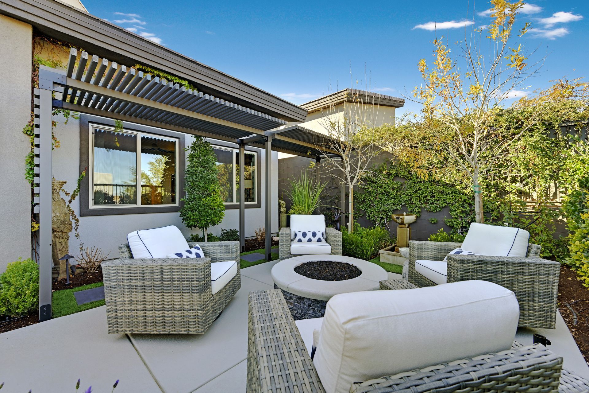 Outdoor patio with seating around a fire pit under a pergola. Green plants and blue sky.