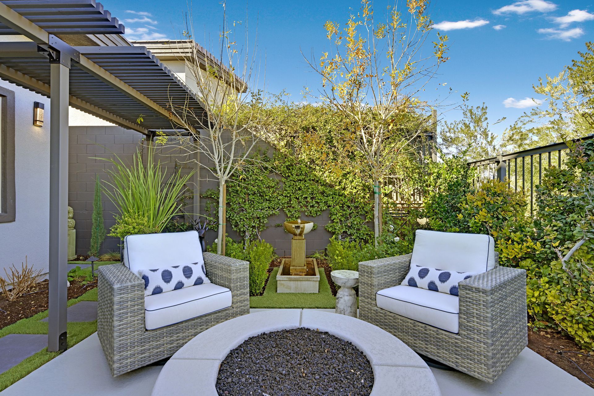 Two wicker chairs with patterned pillows face a fire pit in a small patio garden with a water feature.
