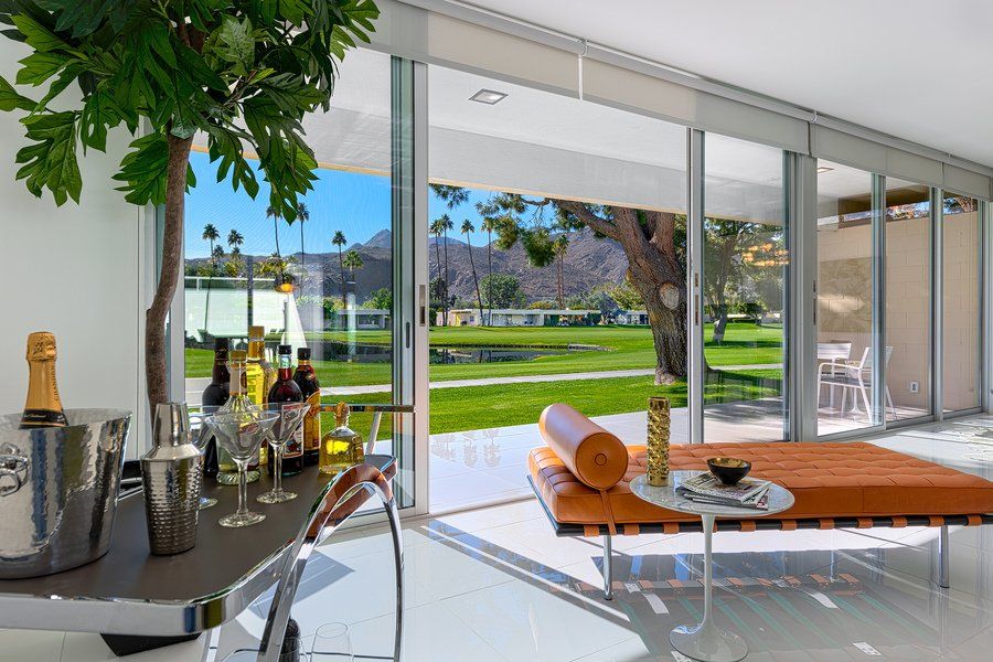 Modern living room with large glass windows, outdoor views, bar cart, and orange chaise lounge.