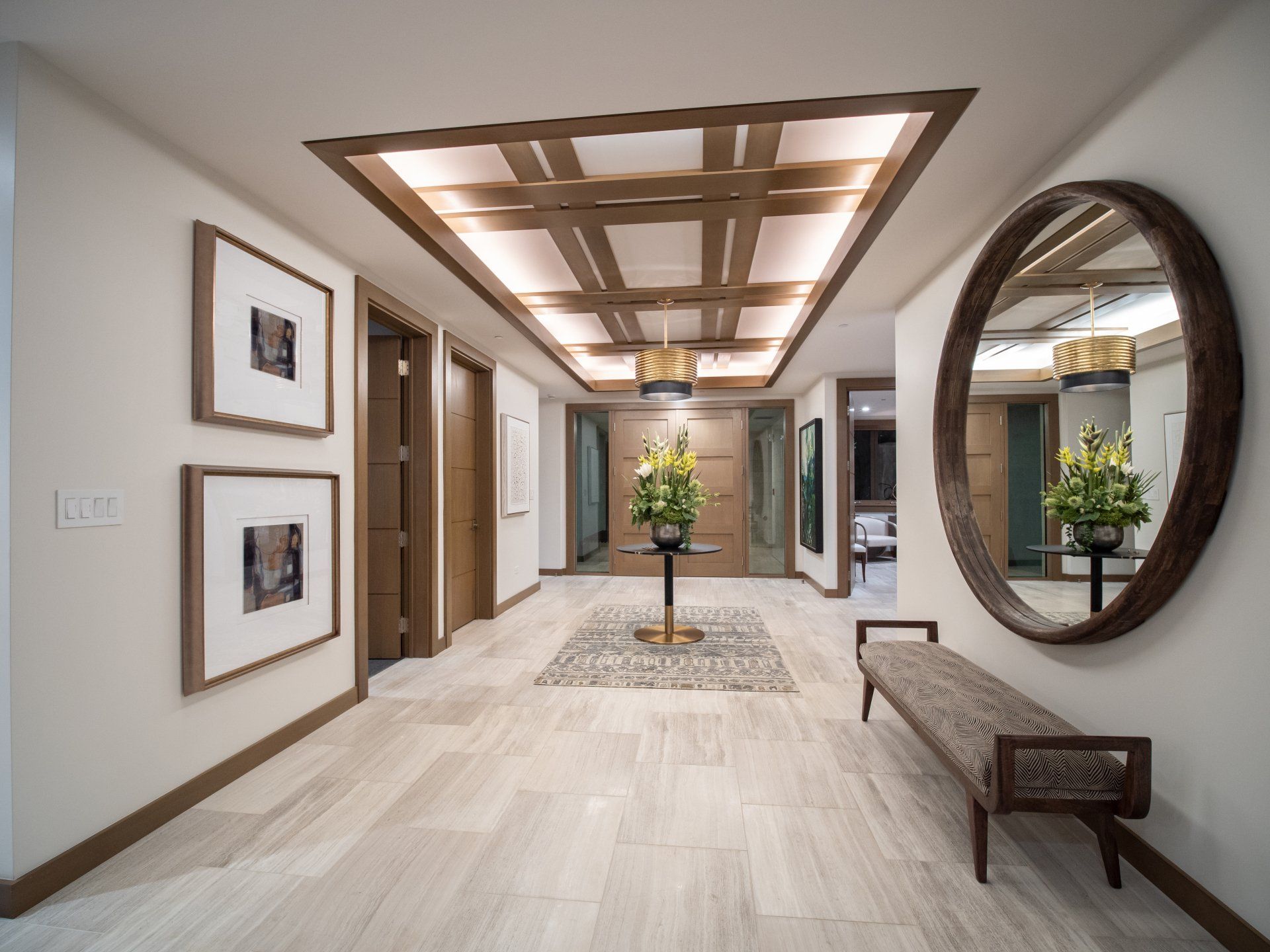 Hallway with light-colored tile floor, wooden ceiling grid, two framed photos, large mirror, and bench.