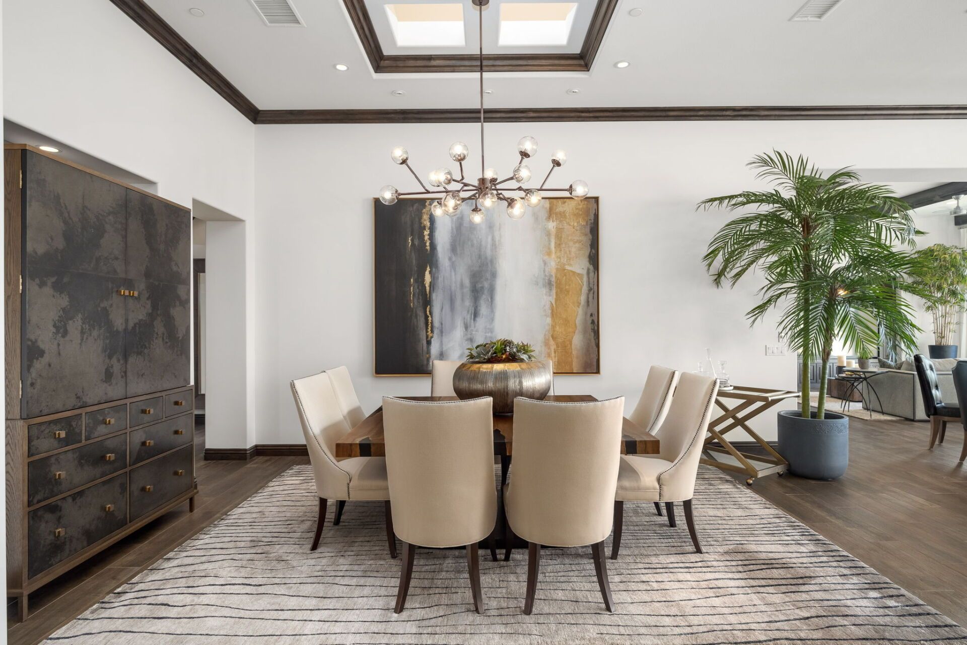 Elegant dining room with table, chairs, art, cabinet, and plant; neutral colors with dark wood accents.