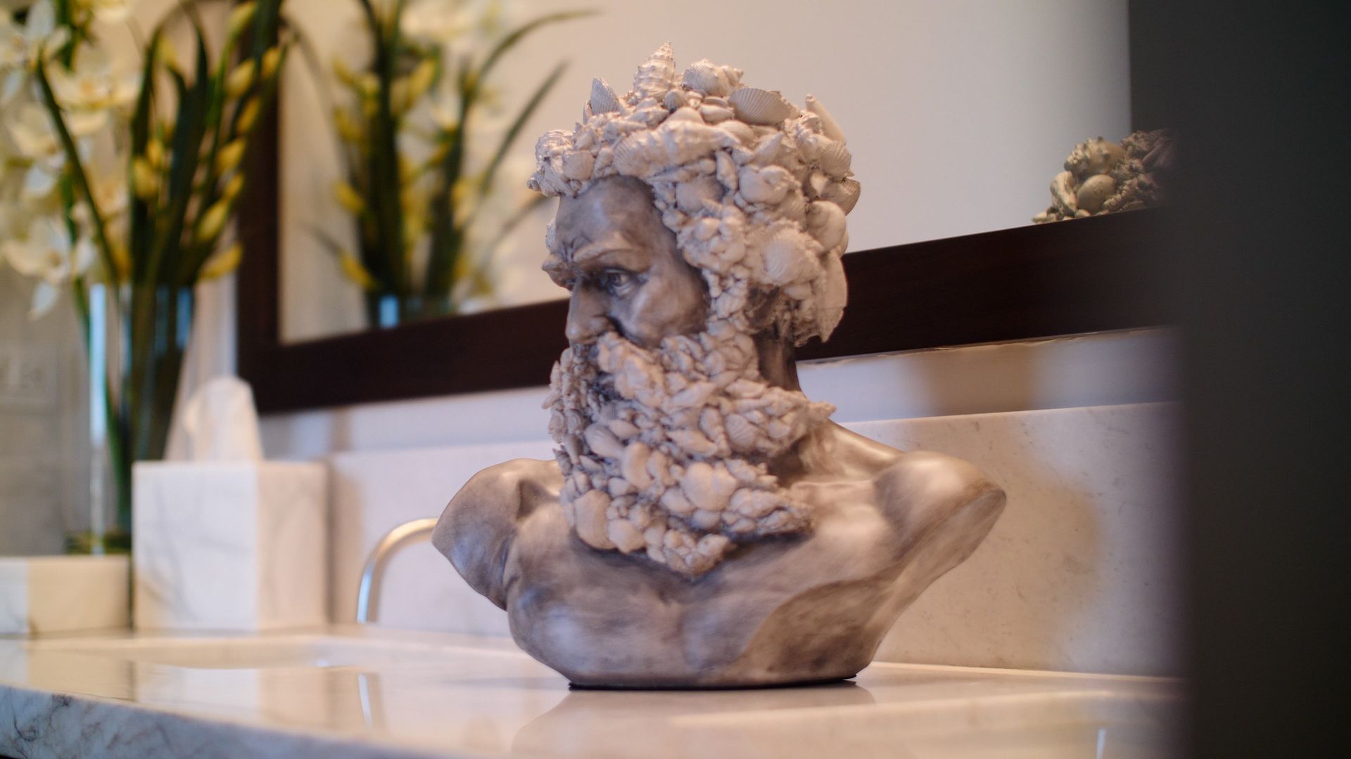 Bust of a bearded man on a bathroom counter, with mirror reflection and vase of flowers.