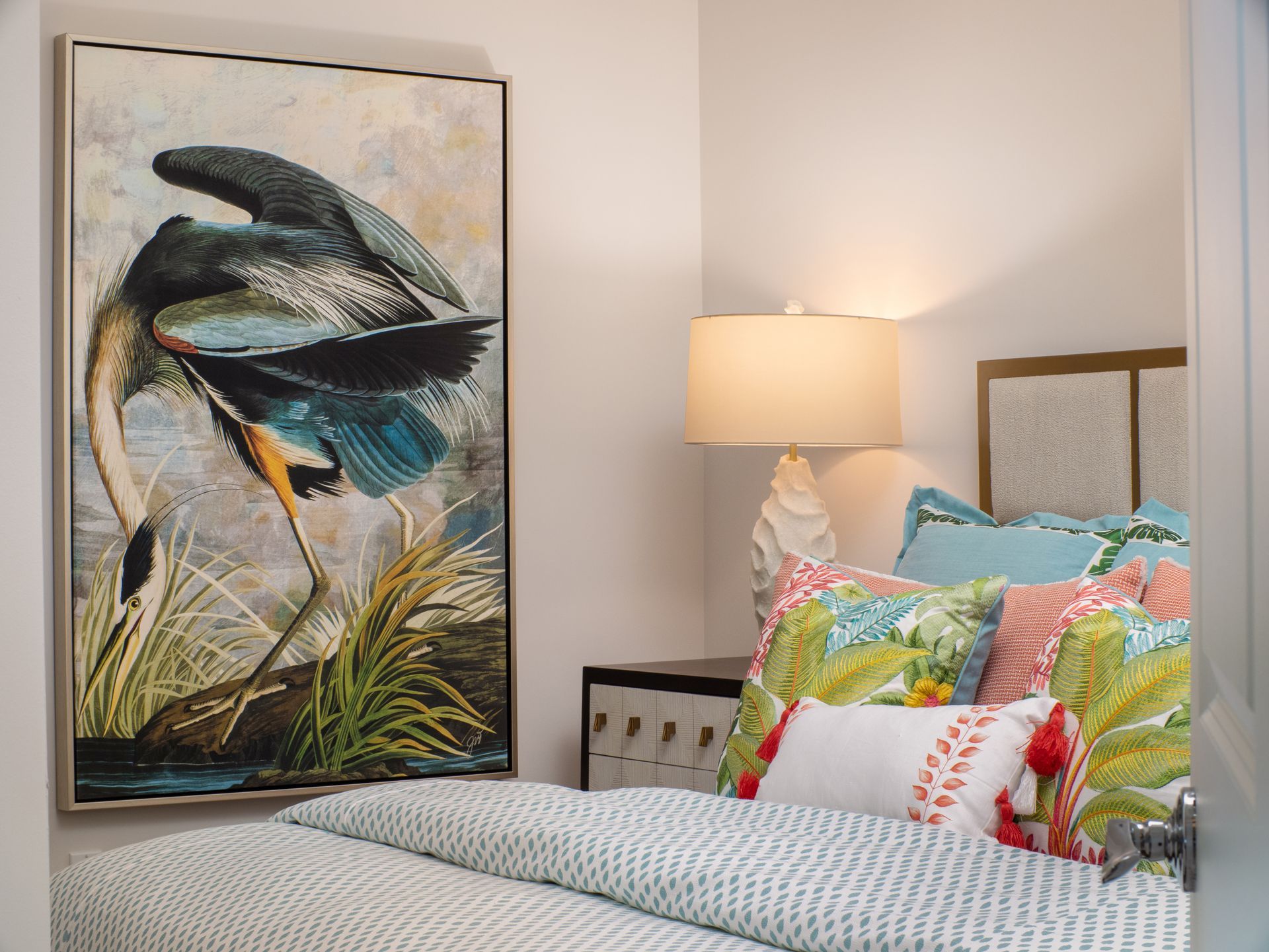 Bedroom with heron painting over a bed with colorful pillows and a bedside lamp.