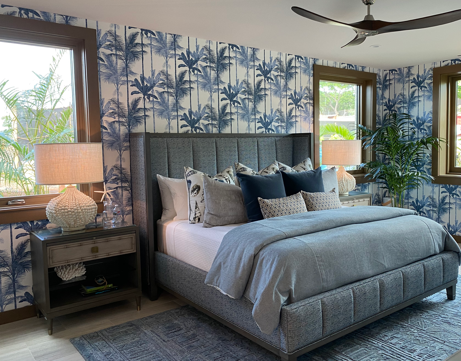 Bedroom with blue palm print wallpaper, gray upholstered bed, and natural light.