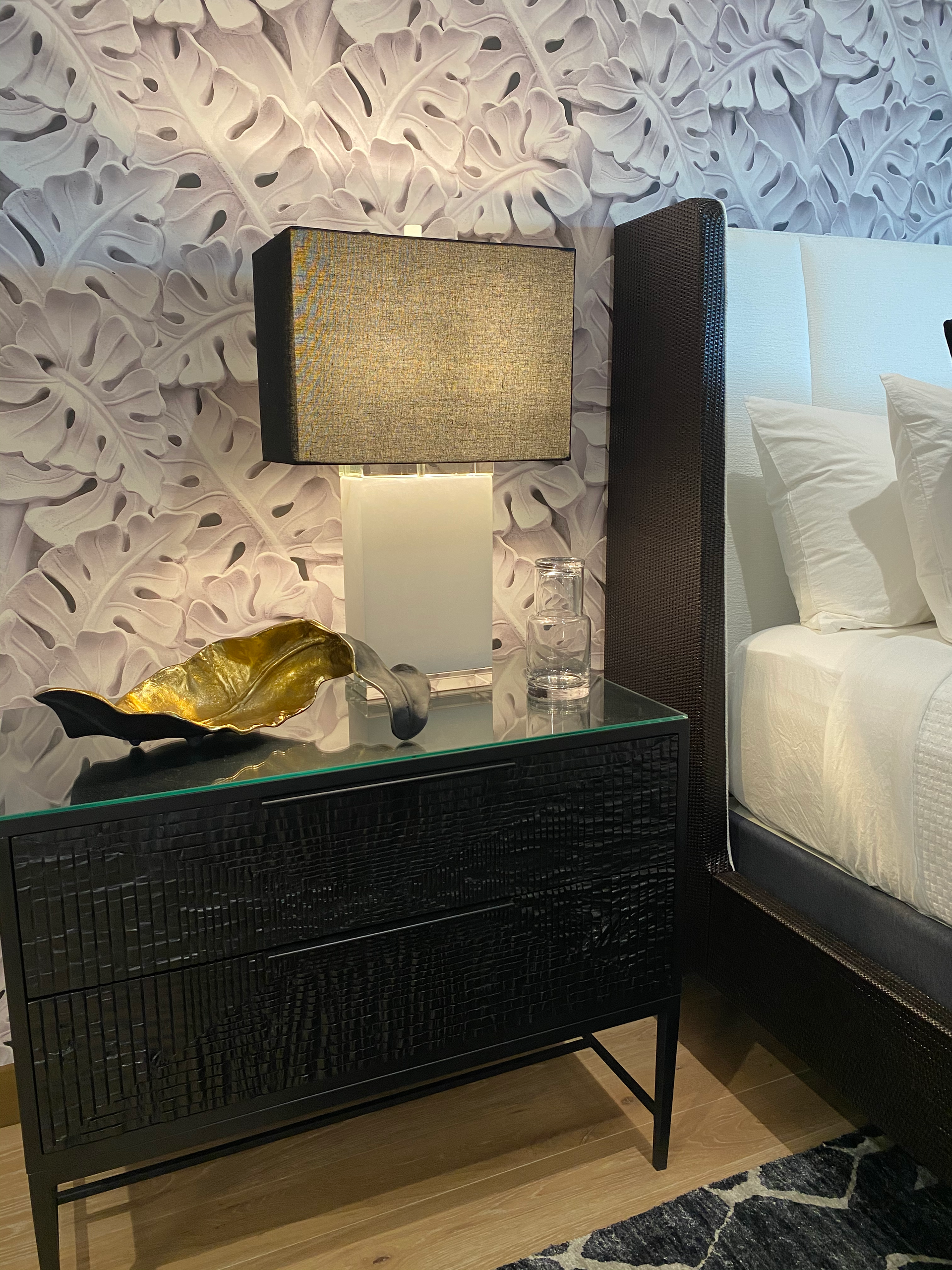 Black nightstand with a lamp, gold bowl, and glass items next to a bed with white linens.