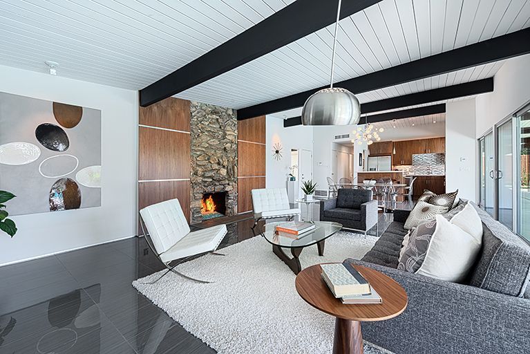 Mid-century modern living room: stone fireplace, white chairs, gray sofa, glass coffee table, wood paneling, and a white rug.