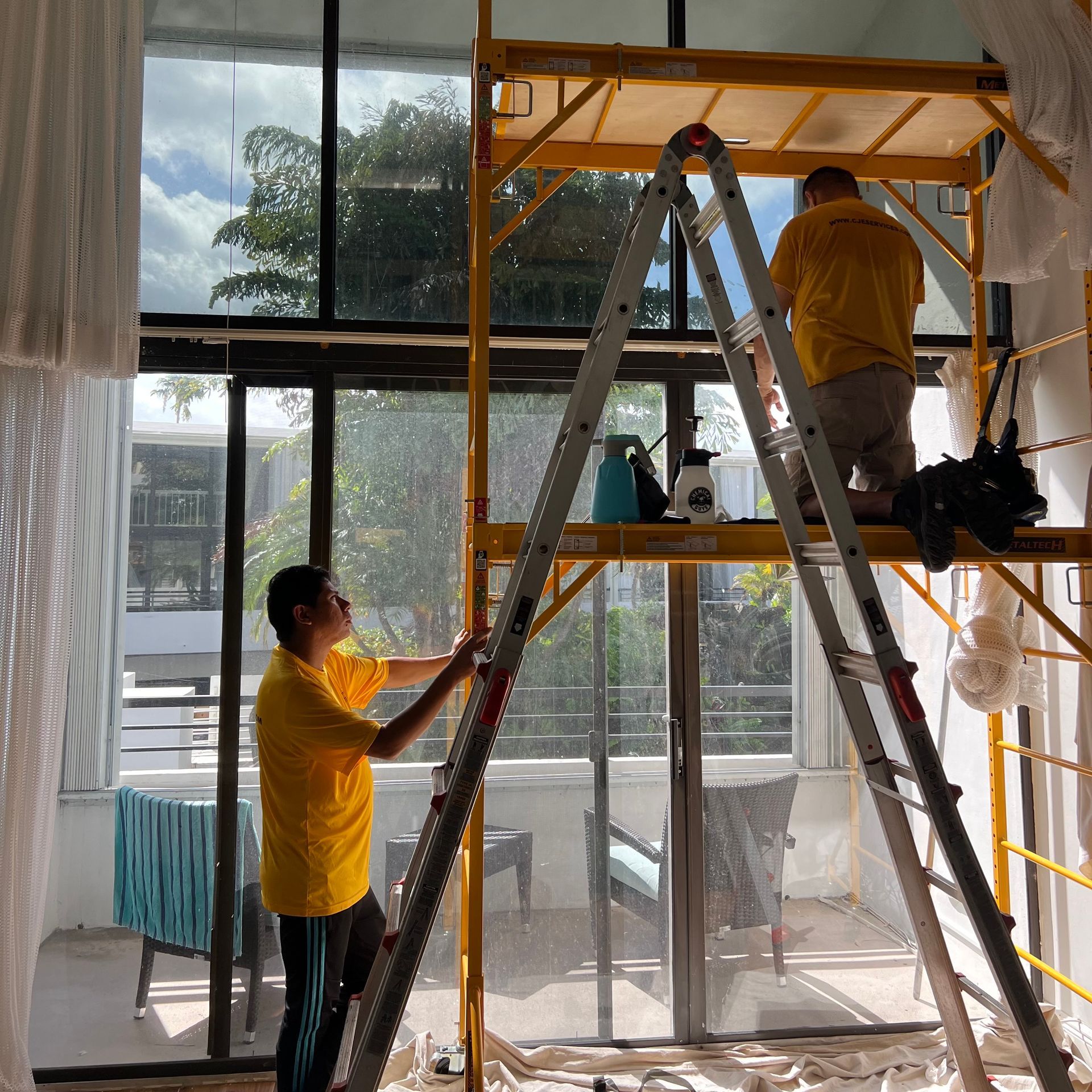 A man in a yellow shirt is standing on a ladder in front of a window.