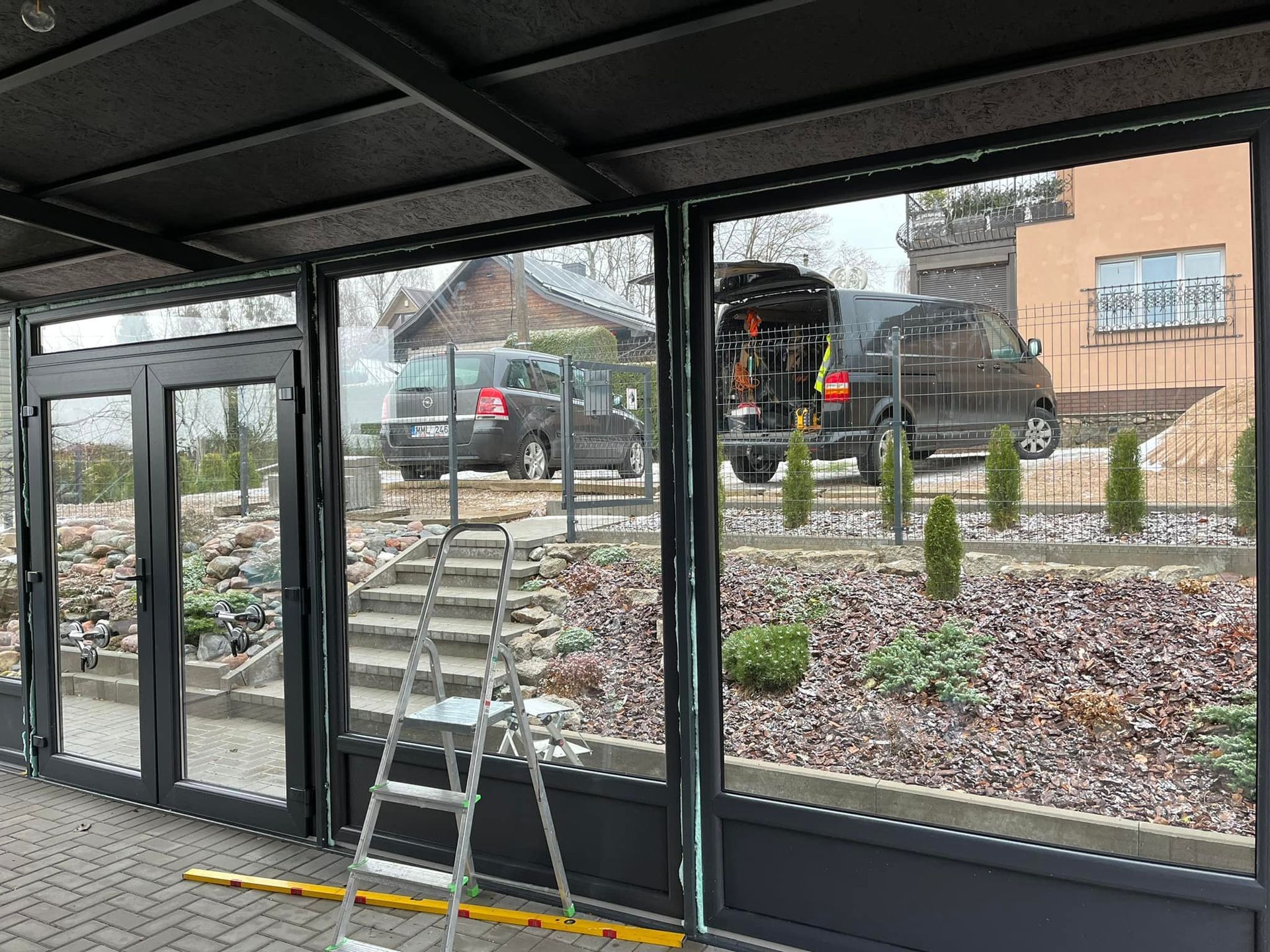 Kopėčios stovi priešais pastatą, kurio fone stovi automobilis.