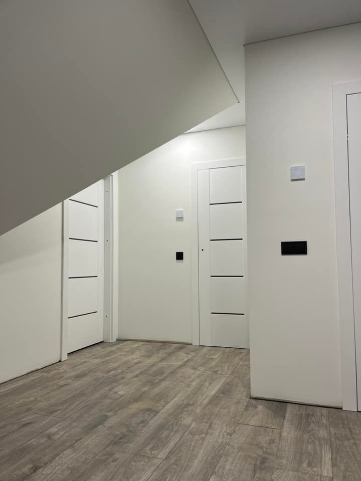 Prieškambaris su baltomis sienomis ir medinėmis grindimis bei laiptais fone.