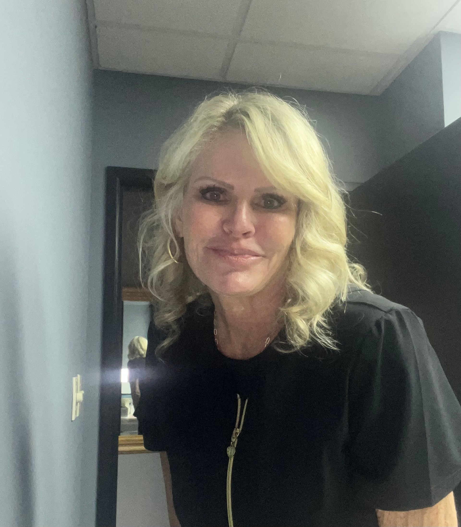 Woman with blonde wavy hair, wearing a black shirt, smiles. Standing in a doorway.