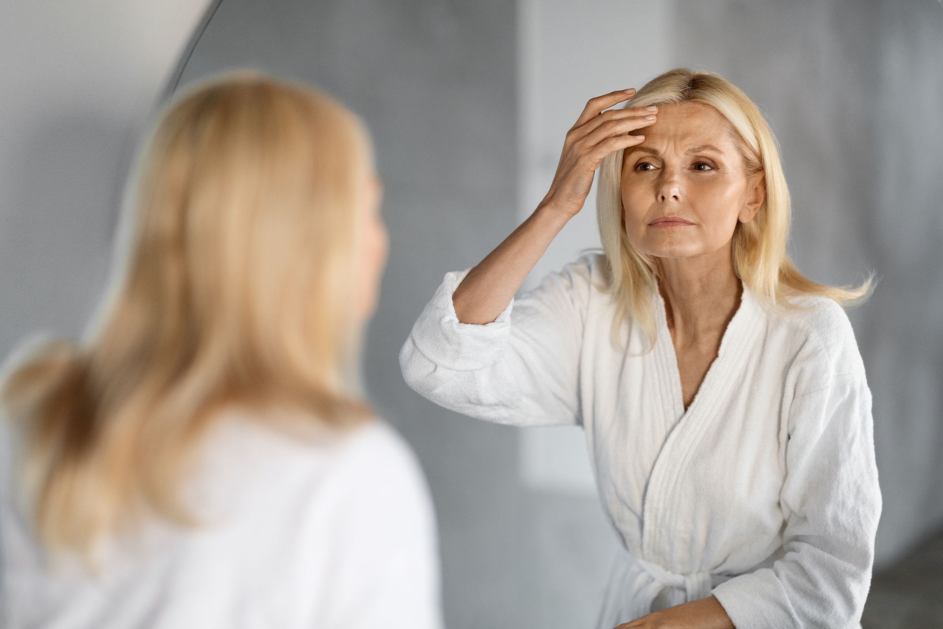 Woman in a white robe looking in a mirror, touching her forehead.