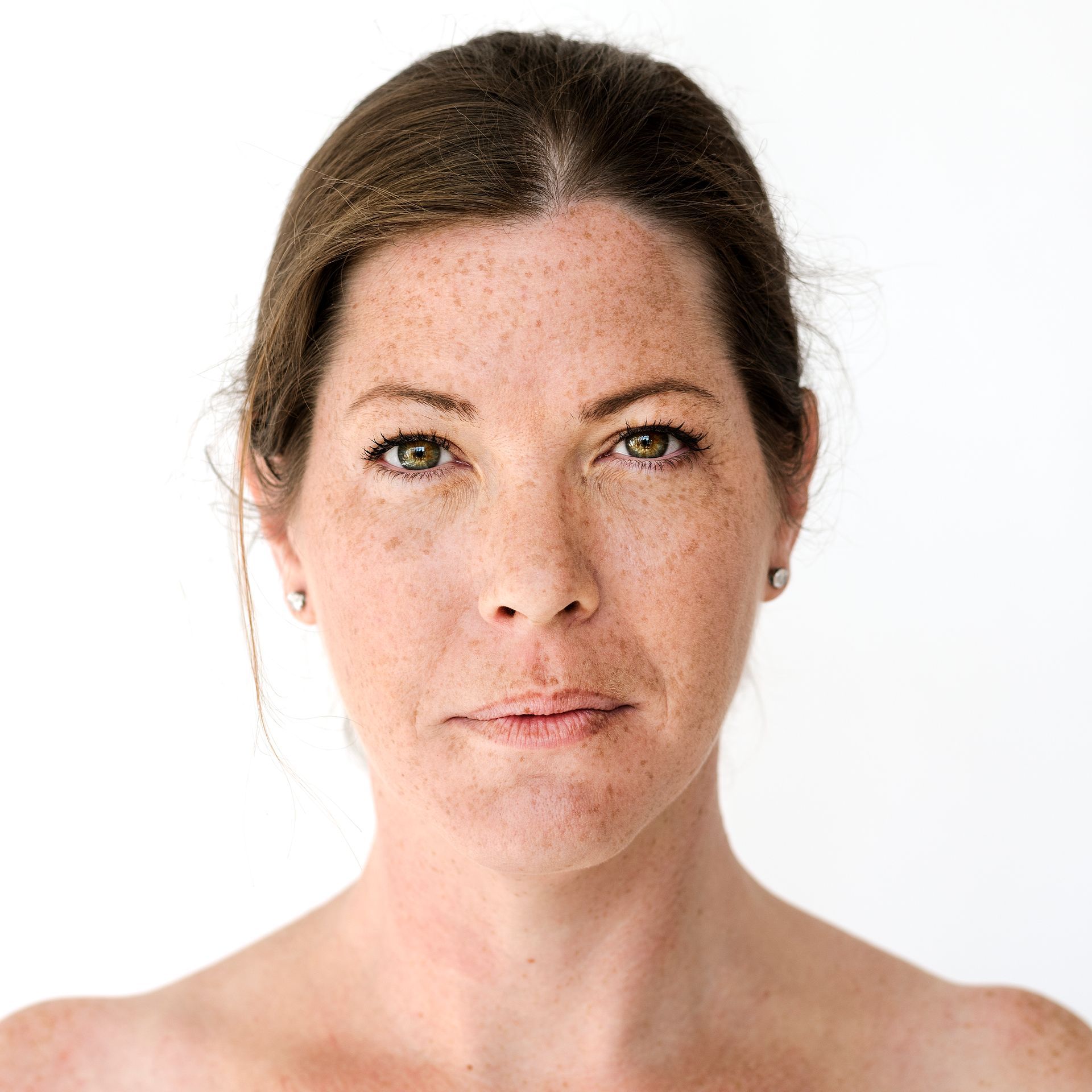 Woman with freckled skin and green eyes, looking directly at the viewer with a neutral expression, bare shoulders, plain white background.