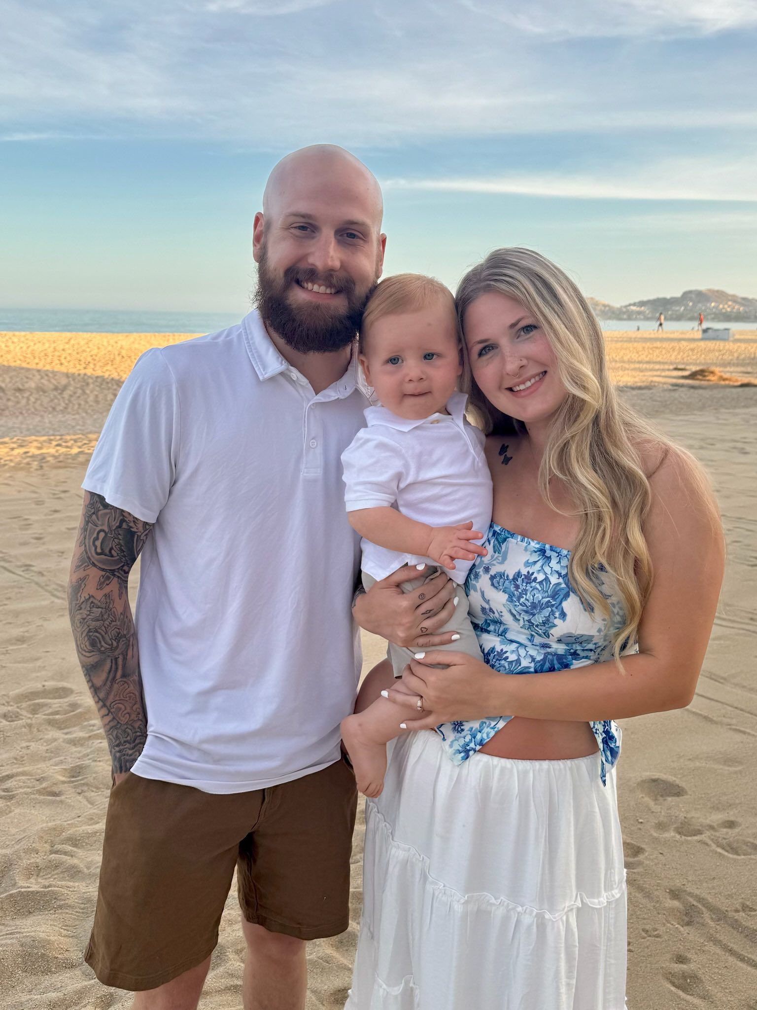 Family of three on beach, man holding baby, woman beside them; smiling, sunny, ocean backdrop.