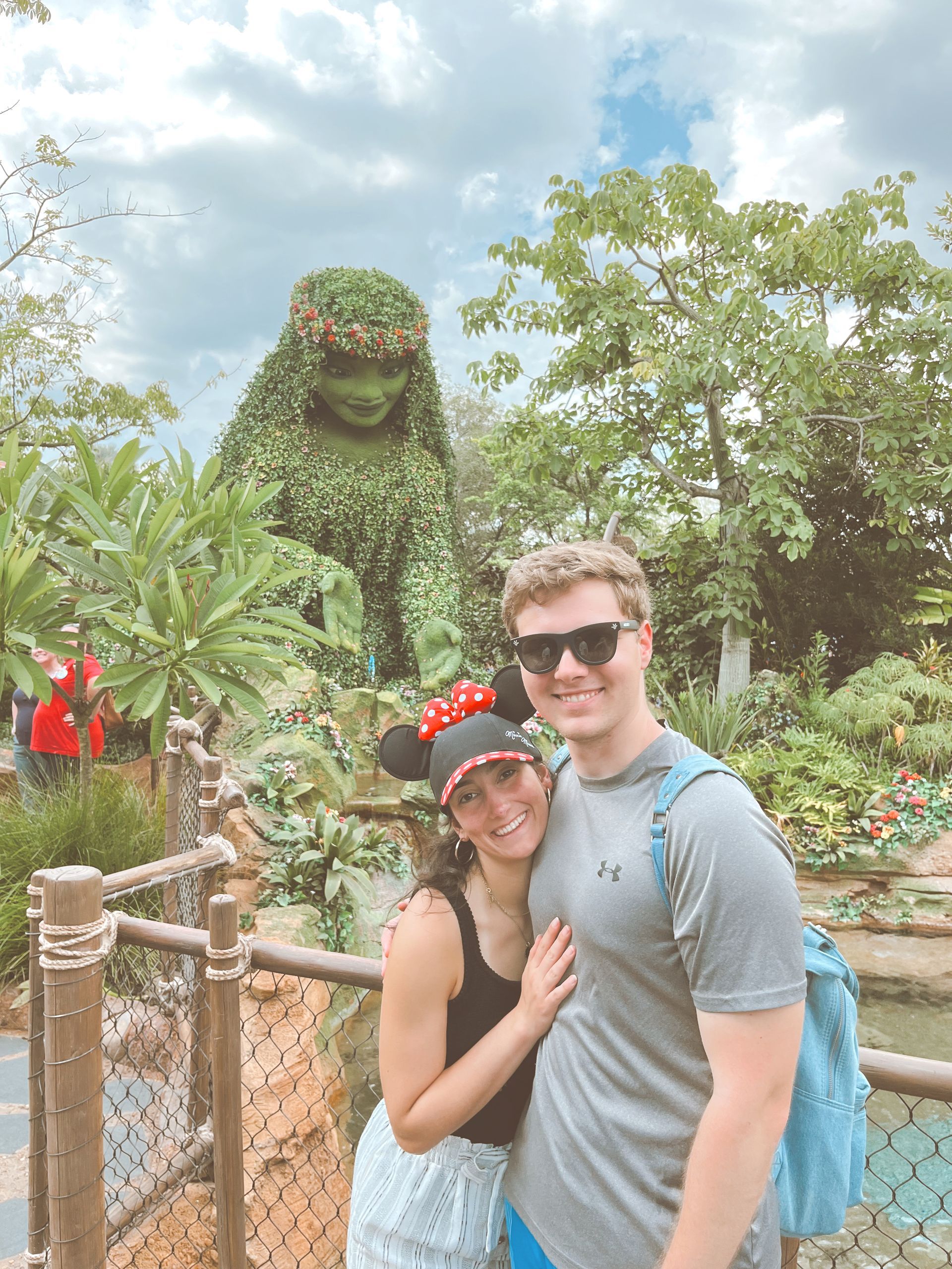 Couple poses in front of a topiary at Disney World. Woman wears Minnie Mouse ears, man a backpack. Sunny day.