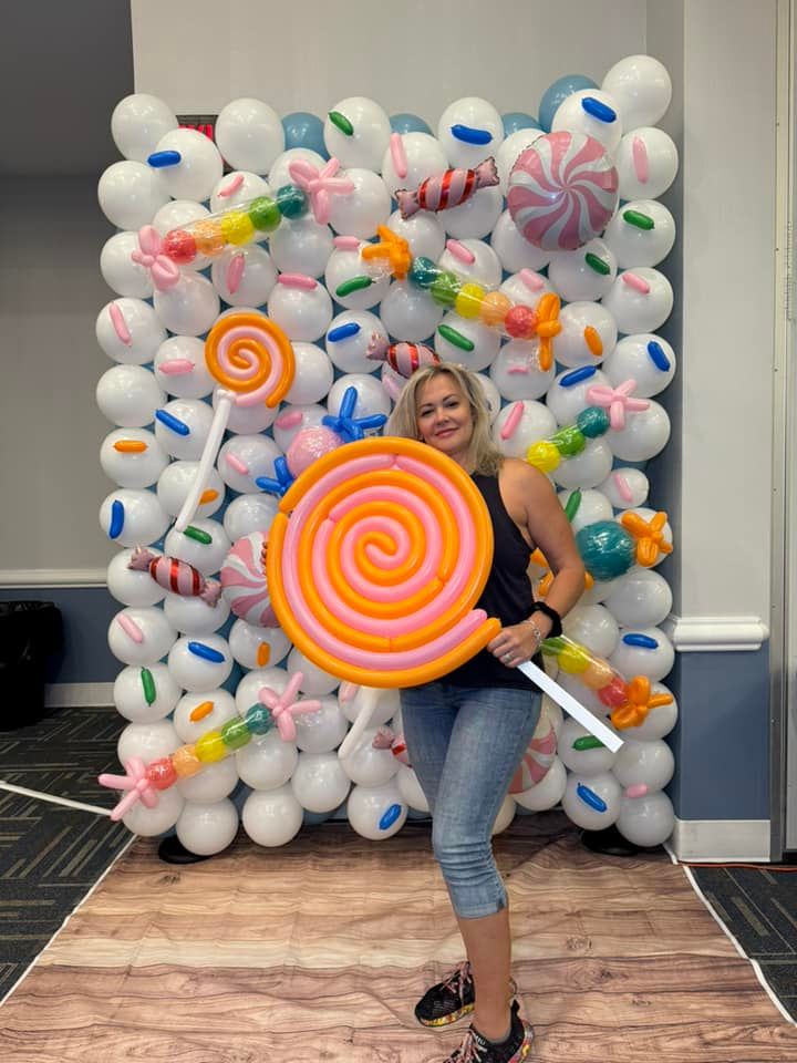 A woman is holding a lollipop in front of a wall made of balloons.